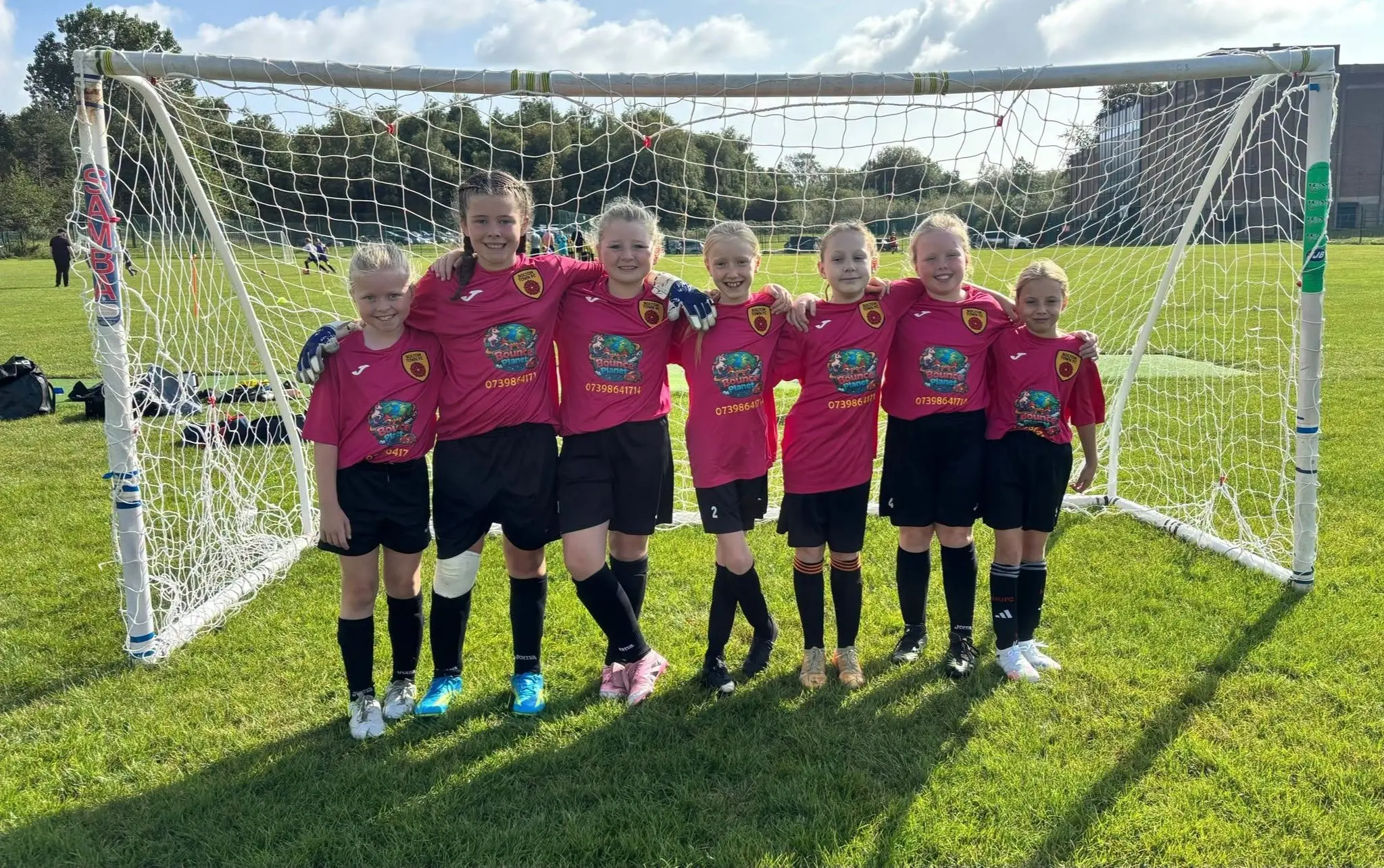 Bolton Town girls in pink kits together in front of their goal on a sunny matchday