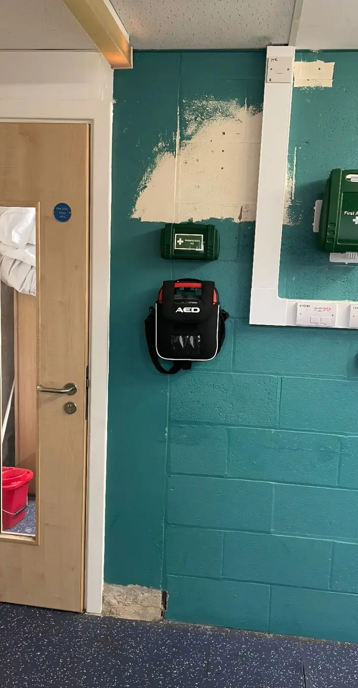 AED mounted on the teal wall beside the first-aid equipment at Health Hub reception
